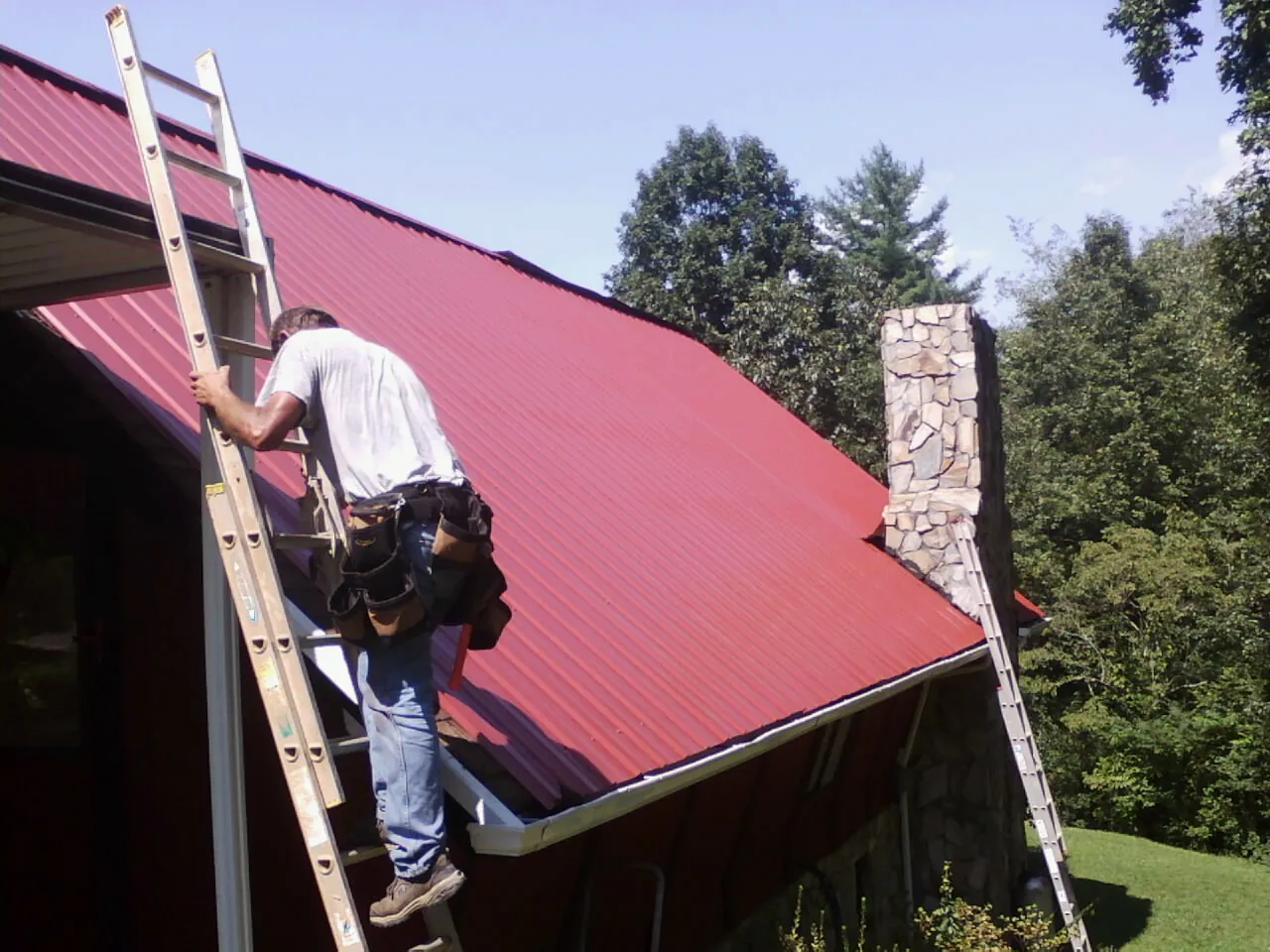 Roofer on a ladder beside a red metal roof and stone chimney — Soffit & Fascia Repair by Roofing Brothers in Dickson